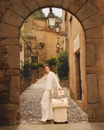 Model standing on a cobblestone street beneath a stone archway, holding a rolling luggage set.