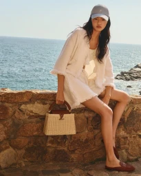Model sitting on a stone wall by the ocean wearing a white tweed jacket, cap, and holding a woven handbag.