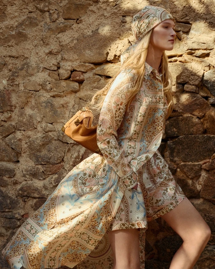 Model in patterned floral print dress and matching headscarf carrying tan leather handbag against a rustic stone wall