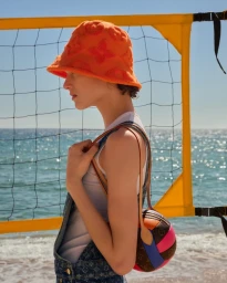 Model in profile wearing an orange bucket hat and denim overalls beside a volleyball net on a sunny beach.