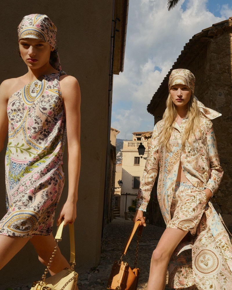 Two models in patterned dresses and matching headscarves walking down a sunny narrow street for Louis Vuitton Resort 2026