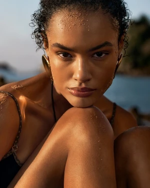 Close-up portrait of a woman with dark curly hair and sand on her forehead, wearing large gold earrings outdoors near water