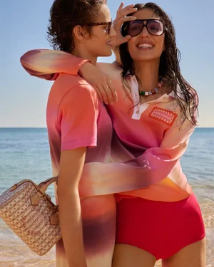 Two smiling women in gradient tops embracing on a sunny sandy beach with the ocean in the background