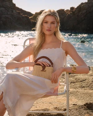 Model seated on a deck chair by the ocean holding a woven straw handbag for the Louis Vuitton Resort 2026 campaign.