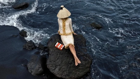 Model wearing a tweed skirt suit and straw hat seated on a dark rock surrounded by churning ocean waves