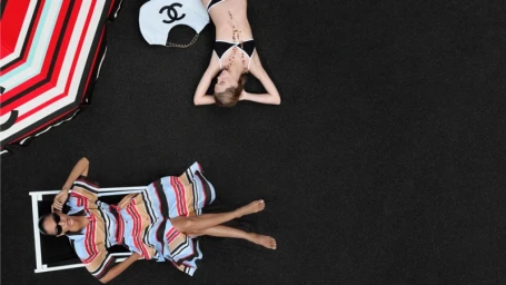 Overhead view of two models lying on a dark surface next to a striped beach umbrella and a white Chanel handbag.