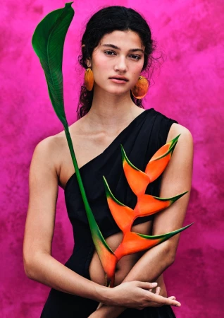 A woman in a black one-shoulder dress holds a vibrant orange heliconia plant against a textured bright magenta background.