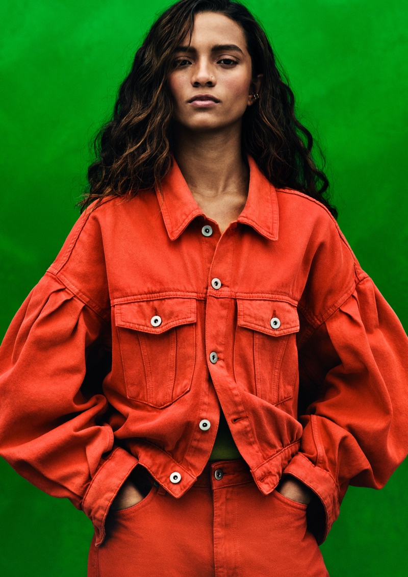 Model wearing a bright orange-red puff-sleeve denim jacket and matching trousers against a vivid green backdrop.