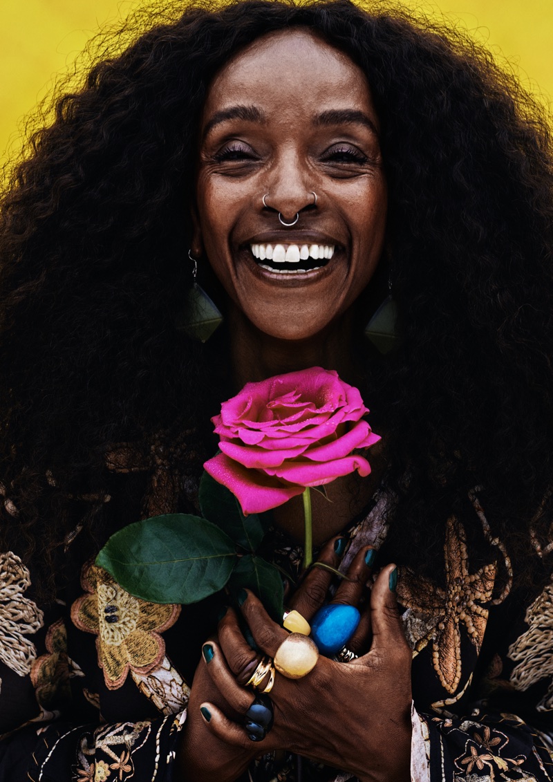 A smiling woman with voluminous dark curly hair holds a bright pink rose against a vibrant yellow backdrop.