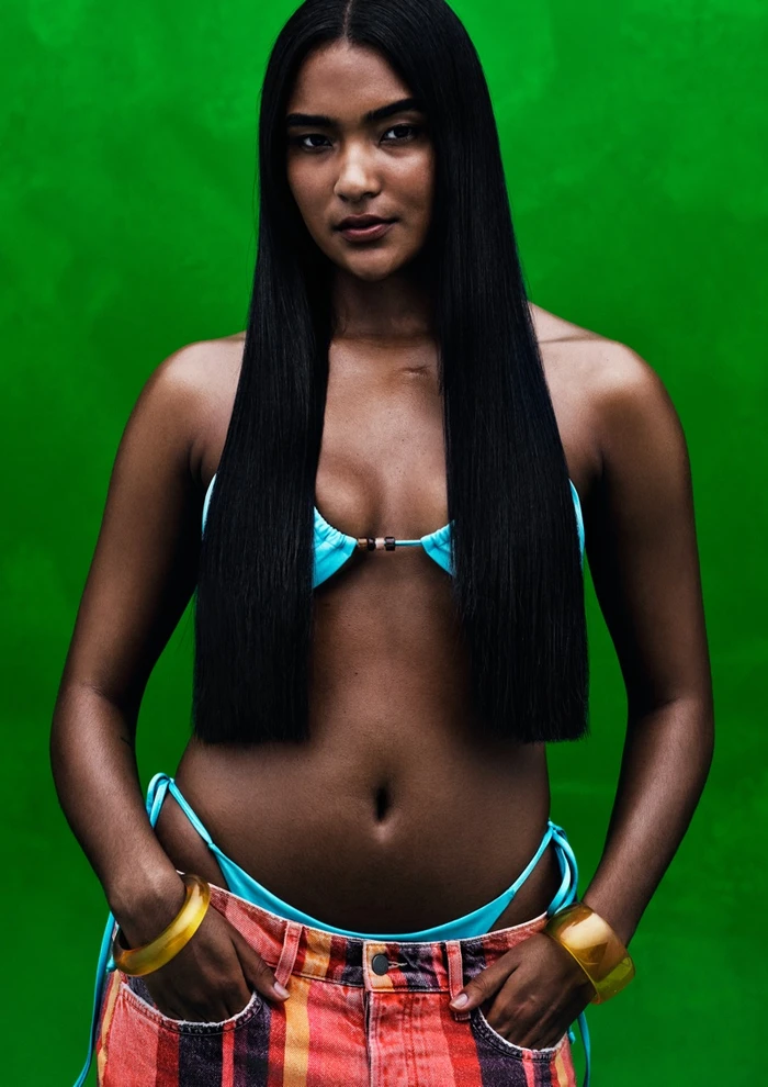 Woman with long black hair wearing a turquoise bikini top and striped trousers against a vivid green background