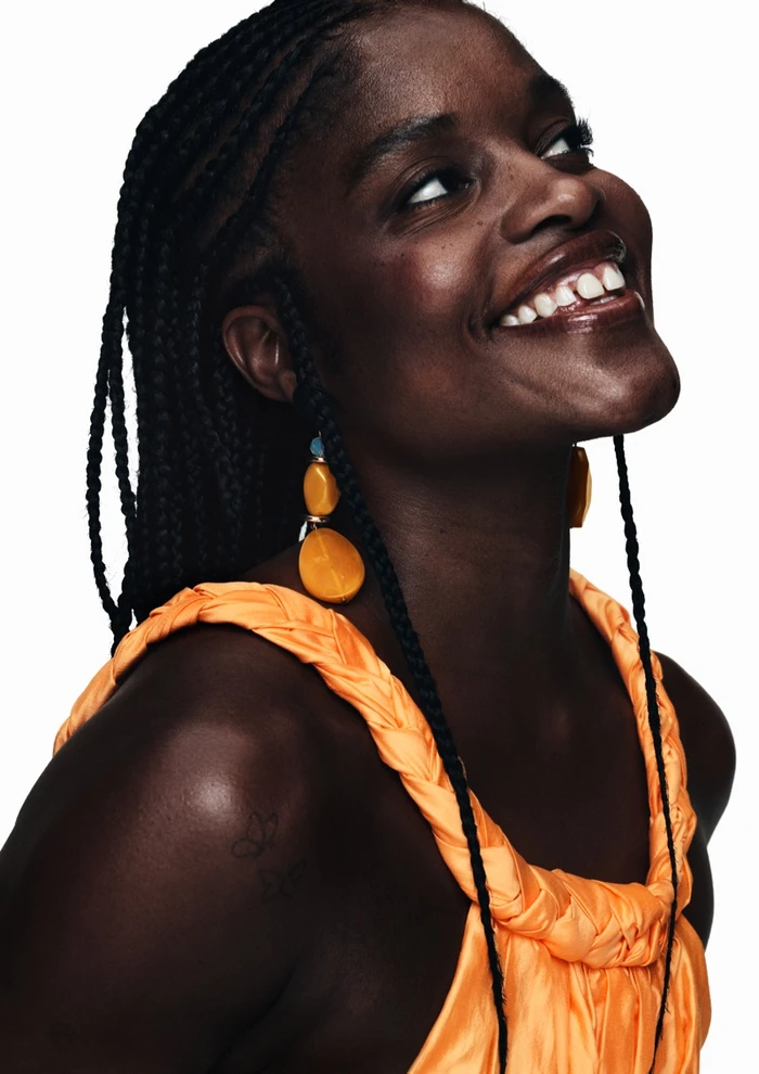 Portrait of Ice Spice in an orange dress wearing large yellow earrings against a plain white background