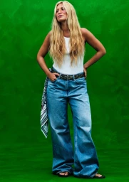 Full-length portrait of a woman with long blonde hair wearing a white tank top and blue denim jeans against a textured green background.