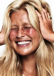 Close-up portrait of a woman with blonde hair, smiling with closed eyes and holding her hands to her head against a plain white background.