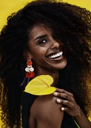 Portrait of smiling woman with voluminous dark curly hair holding a bright yellow flower against a warm background.