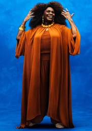 Woman wearing an orange flowing cape and matching trousers posing against a vibrant blue textured studio background.