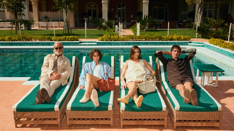 Four models lounging on green-cushioned wicker sunbeds by a swimming pool wearing casual summer attire and loafers.