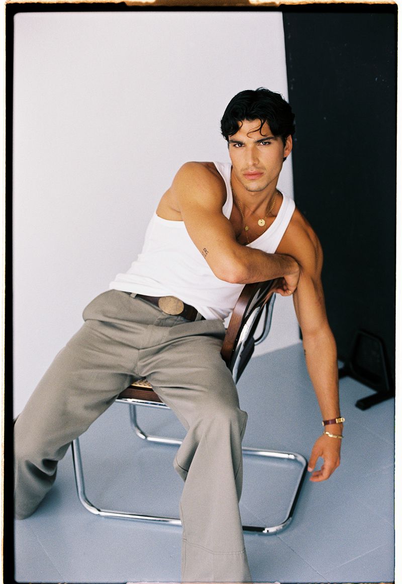 Model Antonio Lopez wearing a white tank top and wide-leg grey trousers seated on a chrome chair in a studio setting.