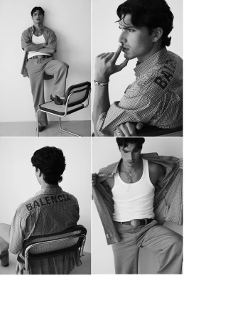 A black and white fashion shot of a young male model in an unbuttoned shirt over a tank top, pants, and dress shoes sitting on a chair.
