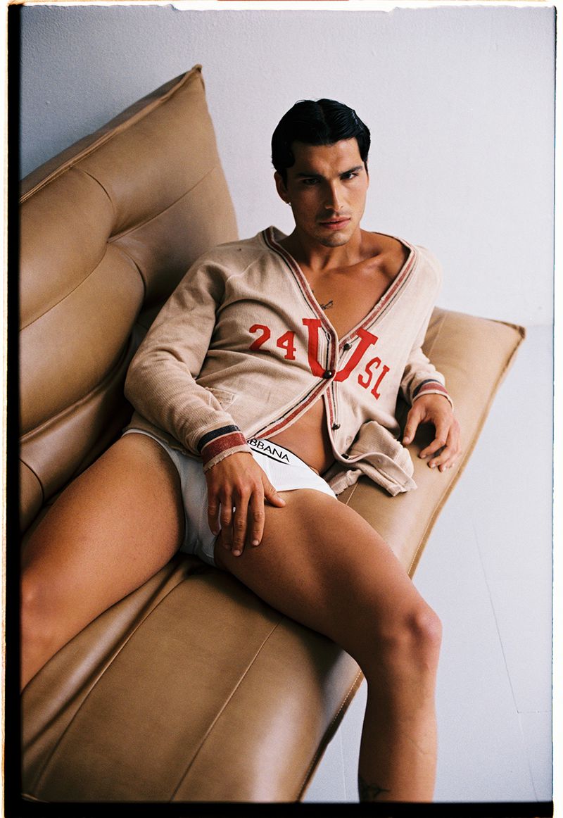 Fashion shot of a male model in a cardigan and underwear, reclining on a brown leather sofa in a bright, simple studio setting.