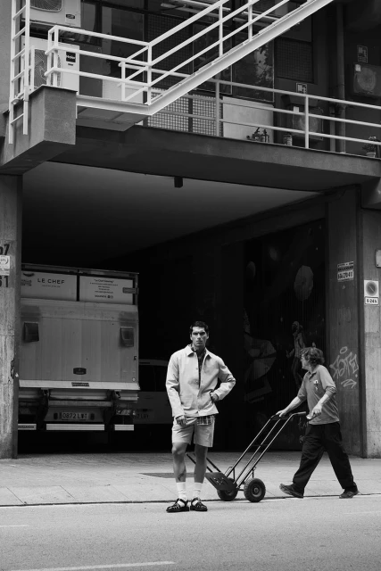 Monochrome shot of two men outside, one standing with a handcart and the other pushing it on a city street near a loading dock.