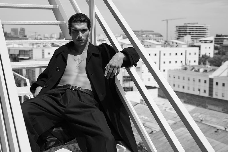Model Antonio Lopez poses shirtless in an open black coat on a white metal rooftop staircase overlooking city buildings in black and white.