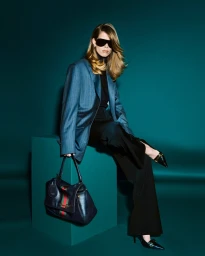 Model wearing dark sunglasses, blue pinstripe blazer, and black trousers while holding a dark leather shoulder bag against a teal studio backdrop.