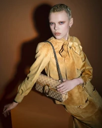 Model with short blond hair wearing a yellow suede bow blouse and holding a monogram handbag against a warm brown wall.