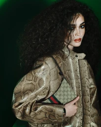 Model with voluminous dark curly hair wearing a snakeskin jacket and holding a monogram handbag for Gucci