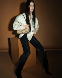 Model with long dark hair wearing a white zip-up jacket and black boots sitting on a box against a brown backdrop