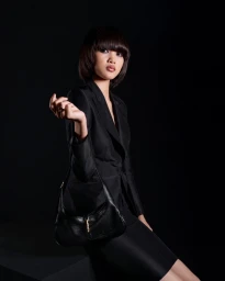 Model with dark bob haircut wearing black blazer holding a black leather handbag against a dark background