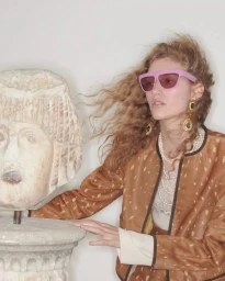 Model Apolline Rocco Fohrer wearing pink sunglasses and brown patterned jacket next to a classical stone bust statue for Valentino