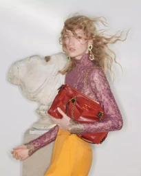 Model with blowing blonde hair wearing purple lace top and holding red handbag against white background
