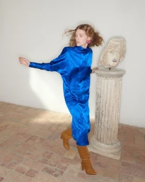 Model wearing a long blue satin dress and brown boots posing near a classical stone pillar and bust