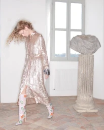 Model with long curly hair wearing a shimmering metallic dress and floral boots walking in a room with a stone pillar.