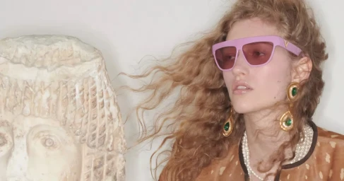 Model Apolline Rocco Fohrer wearing pink sunglasses and ornate earrings next to a stone sculpture for Valentino Pre-Fall 2026.