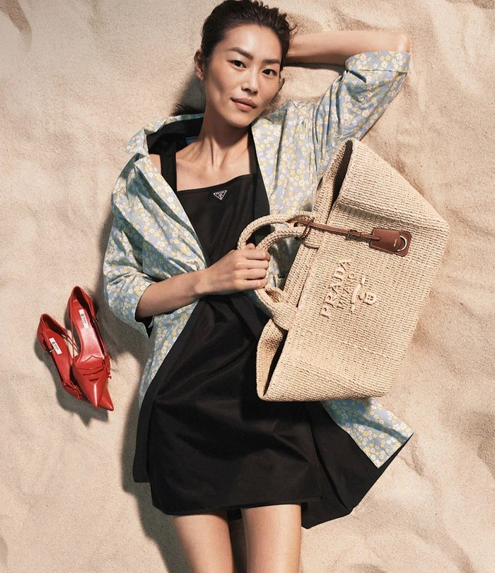 Liu Wen lying on sandy beach holding a woven straw tote bag wearing a black dress and blue floral coat with red pumps beside her