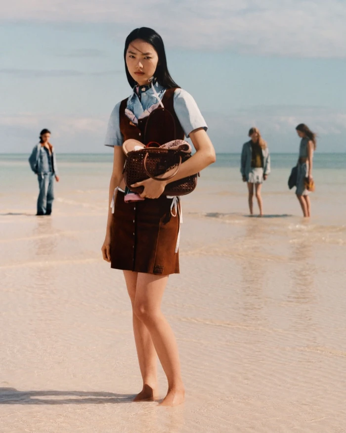 Model standing on a shallow sandy beach holding a dark brown leather handbag, with other figures in the background.