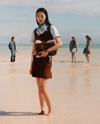 Model standing on a shallow sandy beach holding a dark brown leather handbag, with other figures in the background.