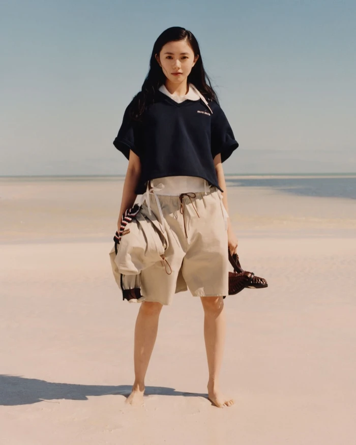 Woman in dark navy polo shirt and beige shorts standing barefoot on a sandy beach for Miu Miu photographed by Jeano Edwards