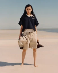 Woman in dark navy polo shirt and beige shorts standing barefoot on a sandy beach for Miu Miu photographed by Jeano Edwards