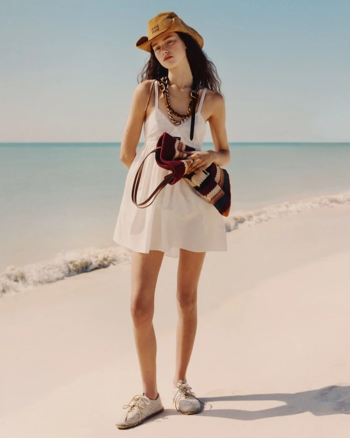 Model in a cream sundress and brown hat holding a striped bag on a sandy beach