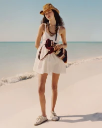 Model in a cream sundress and brown hat holding a striped bag on a sandy beach