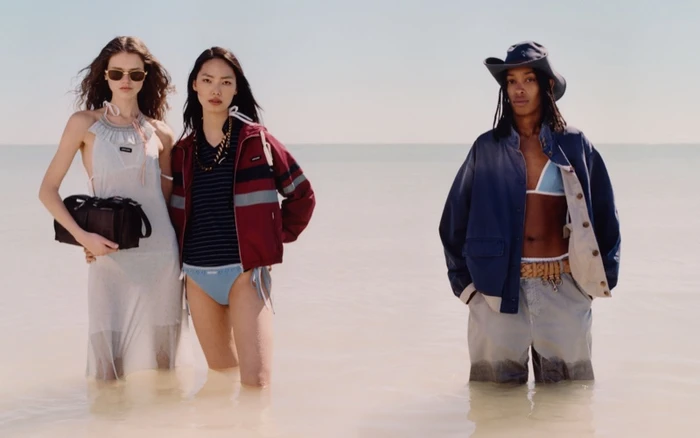 Three models standing in shallow water at a beach wearing resort apparel and outerwear against a clear sky horizon.