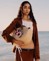 Model holding woven straw bag with floral decoration wearing brown blazer by the ocean for Miu Miu L'Été 2026