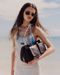Model in sunglasses and light dress holding a dark leather bag against an ocean background for Miu Miu