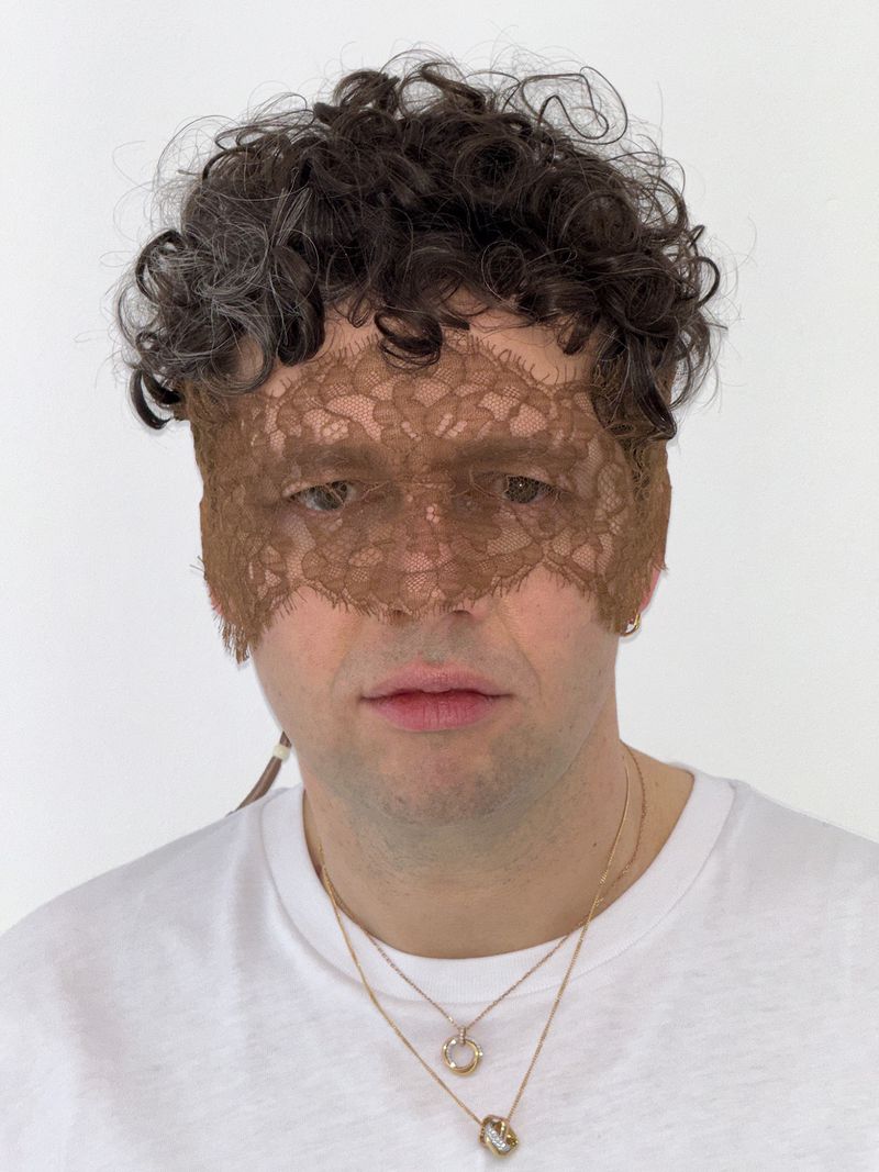 Adult male portrait wearing a lace mask and gold necklaces against a white wall. Shoulder-length curly hair is dark gray.