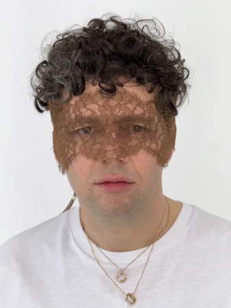 Adult male portrait wearing a lace mask and gold necklaces against a white wall.  Shoulder-length curly hair is dark gray.