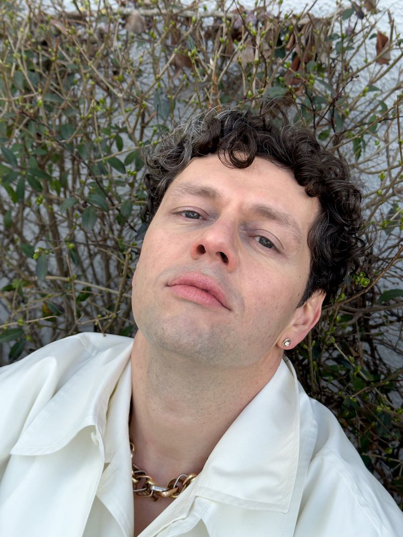Portrait of a curly-haired person wearing a gold chain necklace and a light jacket, posing in front of a bush with budding leaves.