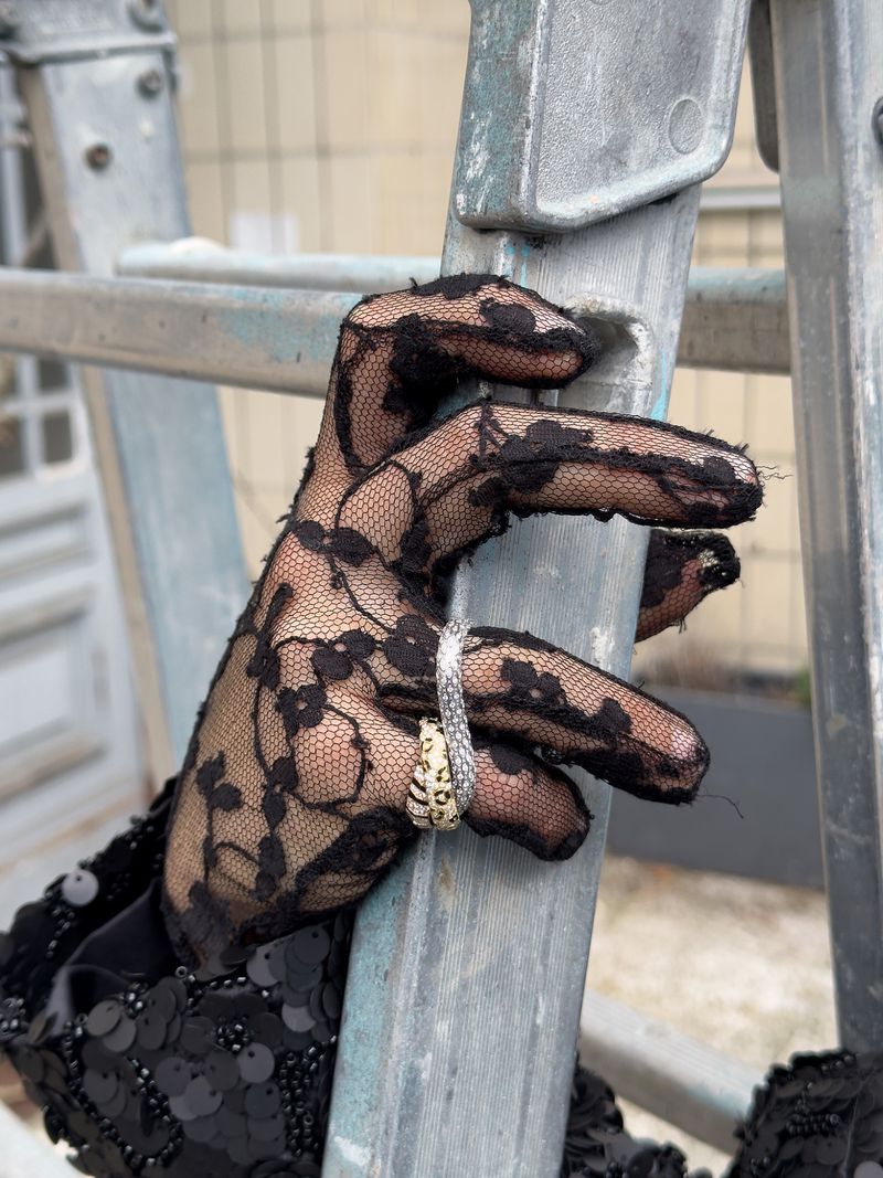A hand wearing a black lace glove holds onto a gray metal ladder, adorned with a ring, set against a blurred outdoor backdrop.