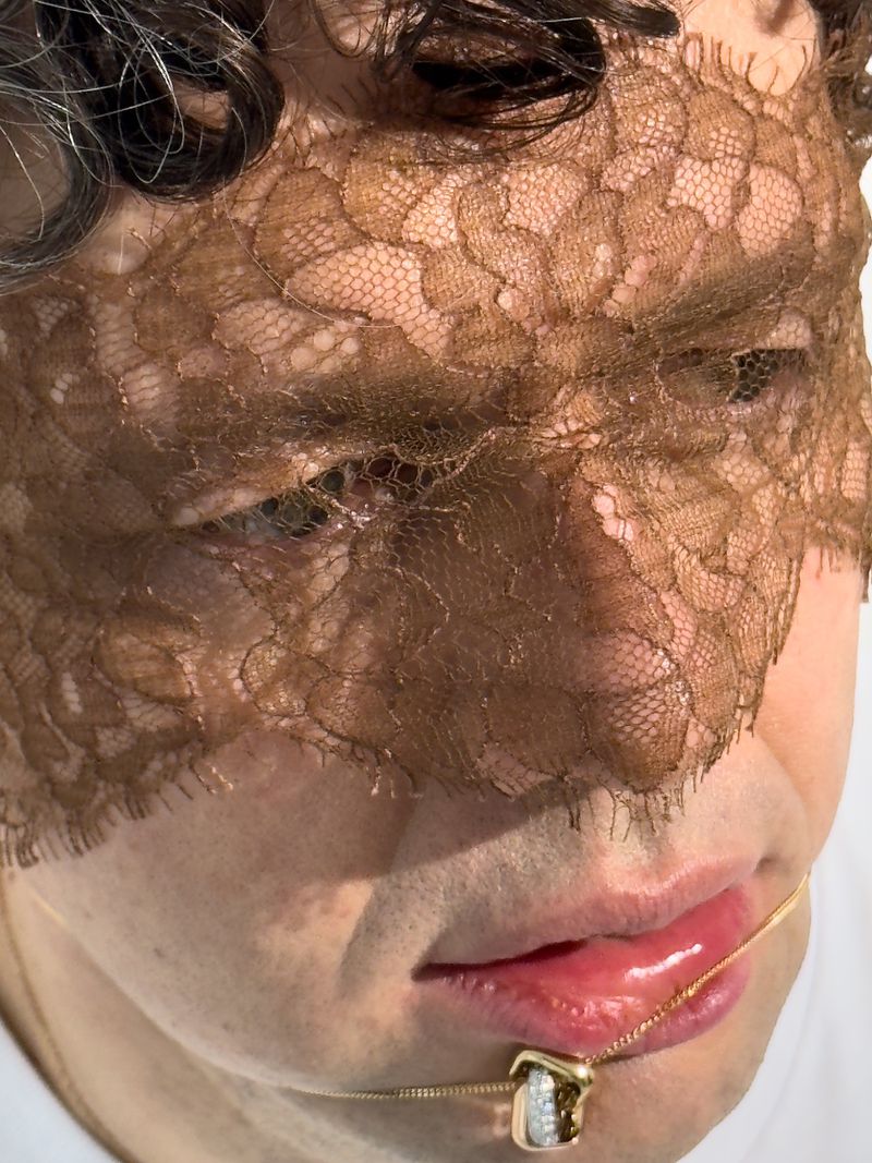 A close-up of a person's face partially covered with lace, emphasizing a gold necklace and soft lighting.